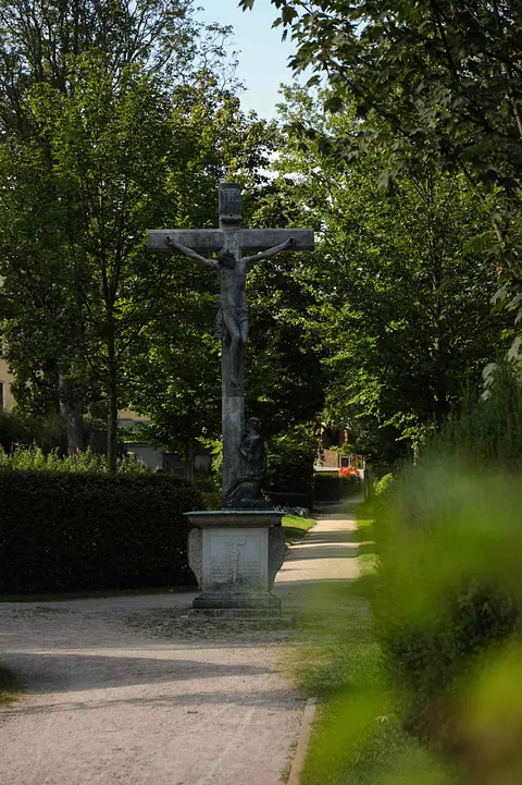 Blick auf das steinere Friedhofskreuz auf dem Friedhof in Schneeberg, rechts und links grüne Laubbäume im Sonnenlicht.